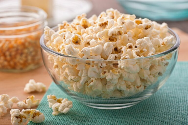 A glass bowl of popcorn with loose kernels beside it